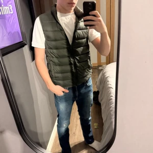 Olivgrön dunväst med ståkrage - Snygg olivgrön dunväst med ståkrage och quiltad design. Västen har dragkedja framtill och är lätt vadderad för extra värme. Perfekt att slänga över en hoodie eller t-shirt för en avslappnad look. Märke och exakt material är okänt.