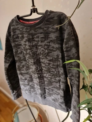 Mörkgrå camo sweatshirt Superdry - Cool mörkgrå sweatshirt från Superdry med camouflagemönster och stor logga framtill. Tröjan har rund halsringning och långa ärmar. Perfekt för en avslappnad streetstyle-look.