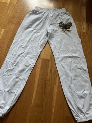 Vita mjukisbyxor med leoparddetalj - Säljer ett par vita mjukisbyxor med resår i midjan och vid bensluten. På ena benet finns en leopardmönstrad mus-öron-detalj och texten 'It's time for dream'. Perfekta för chill dagar och riktigt sköna att ha på sig.