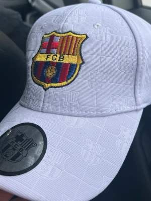 Vit FC Barcelona keps - Snygg vit keps med FC Barcelona-logga broderad framtill och diskret mönster av klubbens emblem över hela kepsen. Justerbar rem baktill och detaljer med texten 'Barcelona' på insidan. Perfekt för dig som älskar fotboll och vill visa ditt stöd för Barça. Skriv gärna för frågor!
