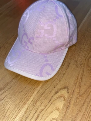 Rosa keps från Gucci med logga - Säljer en snygg rosa keps från Gucci med klassisk böjd skärm och justerbart band bak i beige läder med gulddetalj. Kepsen har tydlig Gucci-logga och är tillverkad i tyg med exklusiv känsla. Perfekt accessoar för dig som gillar lyxiga detaljer.
