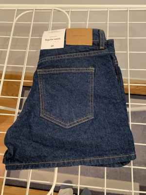 Mörkblå jeansshorts från H&M - Säljer ett par klassiska mörkblå jeansshorts från H&M med regular waist. Shortsen har fem fickor, uppvikta benslut och snygga kontrastsömmar. Perfekta till sommaren och lätta att matcha med olika stilar.