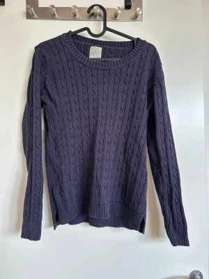 Mörklila kabelstickad tröja - Snygg mörklila kabelstickad tröja med rund halsringning och långa ärmar. Tröjan har ribbade muddar vid ärmslut och nederkant, samt ett klassiskt kabelmönster över hela plagget. Perfekt för lager-på-lager och enkel att matcha med jeans eller kjol.