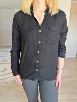 Svart skjorta med knappar - Säljer en svart skjorta med klassisk krage och snygga bruna knappar framtill. Skjortan har två bröstfickor och långa ärmar. Helt oanvänd 