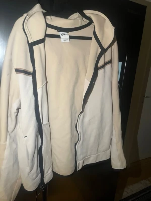 Beige Nike hoodie med dragkedja - Snygg beige hoodie från Nike med hel dragkedja och svart Nike-logga på bröstet. Hoodien har en huva med dragsko, svarta detaljer längs dragkedjan och en tunn rand över bröstet. Perfekt för en avslappnad streetstil.