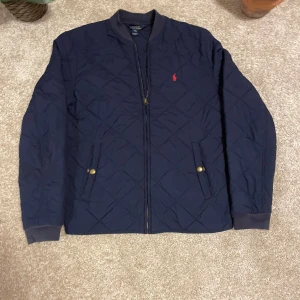 Blå quiltad jacka från Ralph Lauren - Säljer en snygg bli quiltad jacka från Ralph Lauren. Den har en broderad röd logga på bröstet och praktiska fickor med knappar. Perfekt för höst och vår med sin stilrena design och bekväma passform. Dragkedja framtill för enkel stängning.