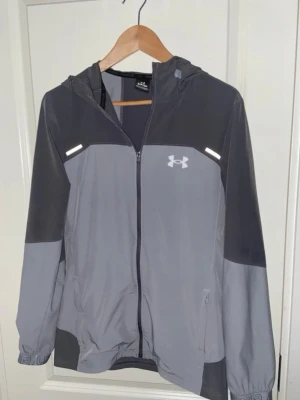 Grå vindjacka från Under Armour - Snygg grå och mörkgrå vindjacka från Under Armour med huva, hel dragkedja och reflekterande detaljer. Jackan har logga på bröstet och stor text på ryggen. Perfekt för blåsiga dagar och sportiga outfits.