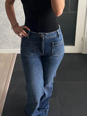 Blå bootcut jeans med fickor fram - Säljer ett par blå bootcut jeans med två stora fickor framtill och hög midja som slutar strax under naveln. Jeansen har en klassisk denimlook och stängs med dragkedja och knapp. Perfekta för en avslappnad och trendig stil. Storlek XS, aldrig använda endast provade 💕💕
