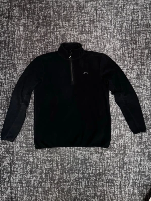  fleece-tröja från Oakley - Svart långärmad fleece-tröja från Oakley med halv dragkedja och hög krage. Tröjan har Oakleys logga broderad på bröstet och är tillverkad i mjukt fleecematerial som håller dig varm under kyliga dagar. Storlek S