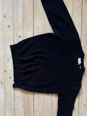 Svart stickad tröja från Les Deux - Klassisk svart stickad tröja från Les Deux med rund halsringning och ribbade muddar. Enkel och stilren design som passar perfekt till jeans eller chinos. Tröjan är mjuk och skön, perfekt för lager-på-lager under kyliga dagar eller som yttertröja mildare vårdagar.