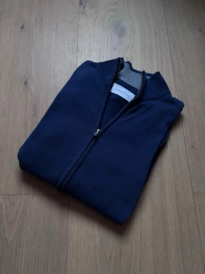 Mörkblå ziptröja från Pier One - Mörkblå ziptröja från Pier One med ribbad krage och dragkedja framtill. Tröjan har en stilren design och är tillverkad i ett mjukt, stickat material. Perfekt för lager-på-lager och enkel att matcha med andra plagg.