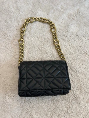 Zara, svart quiltad väska - Snygg svart axelväska i quiltat mönster med guldfärgad kedja som axelrem. Perfekt för dig som vill ha en trendig och stilren accessoar. Aldrig kommit till användning, därav säljer jag den.