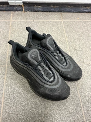Nike Air Max 97 helsvarta sneakers - Nike Air Max 97 sneakers i helsvart med ikoniska vågformade linjer och synlig Air Max-sula. Ovandelen är i textil och syntet, med svarta snören och dragflikar både fram och bak. Perfekta för dig som gillar en clean och sportig look.