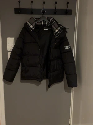 Svart pufferjacka med huva - Snygg svart pufferjacka från Burberry med rutigt foder i huvan och tryck på ärmen. Jackan har en rejäl huva och dragkedja framtill. Perfekt för kalla dagar när du vill hålla stilen. Klassisk design med moderna detaljer.