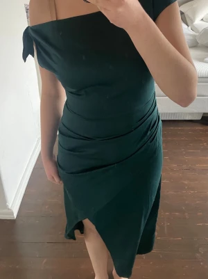 Mörkgrön omlottklänning offshoulder - Snygg mörkgrön omlottklänning med offshoulder och knyt på ena axeln. Klänningen har en asymmetrisk nederkant och draperade detaljer längs midjan. Perfekt för dig som vill ha en elegant och trendig look.