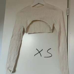 Vit-beige bolero ribbad långärmad - Trendig beige-vit bolero med lång ärm och ribbad struktur. Toppen har en unik öppen front och är perfekt för att styla lager på lager. Tillverkad i ett mjukt material som ger en bekväm känsla och snygg passform.