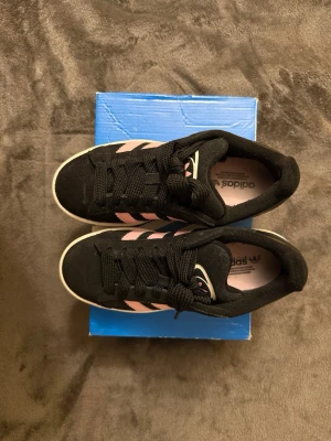 Adidas svartrosa sneakers i mocka - Snygga Adidas sneakers i svart mocka med klassiska rosa ränder på sidorna och vit sula. Skorna har lågt skaft och svart snörning. Perfekta för dig som gillar en sportig och stilren look med en touch av färg.