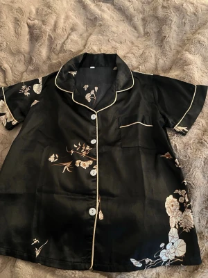 Svart blommig pyjamaströja - Kortärmad pyjamaströja i svart satin med vita och beige blommönster. Tröjan har vit kontrastpiping längs krage, knappslå och ficka samt vita knappar framtill. Perfekt för en chill kväll hemma. Pris kan diskuteras❤️