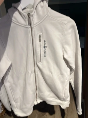 Vit fleecejacka från Sail Racing - Snygg vit fleecejacka från Sail Racing med hel dragkedja framtill och en bröstficka med dragkedja. Jackan har hög krage och huva, samt logga på bröstet. Perfekt för lager-på-lager och chill vardagsstil.
