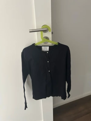 Svart kofta från Zara - Stilren svart kofta från Zara med rund halsringning och knäppning framtill med pärlknappar. Långärmad modell i mjukt material som passar perfekt till lager på lager. Enkel och klassisk design som funkar till det mesta.