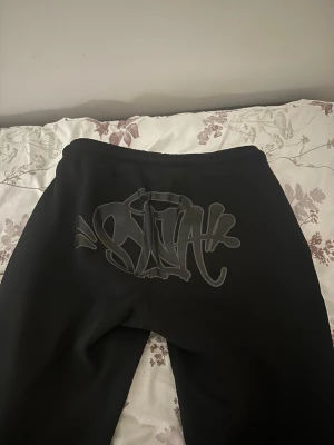 Svarta mjukisbyxor med brodyr - Säljer ett par svarta mjukisbyxor med coolt graffiti-tryck bak och broderad text 'Forgive Me' på benet. Byxorna har avslappnad passform och är gjorda i mjukt material, perfekta för chill dagar. Snygg streetwear vibe! Köpt från ett syna world släpp kvitto finns 100% äkta pris kan diskuteras vid snabb affär 