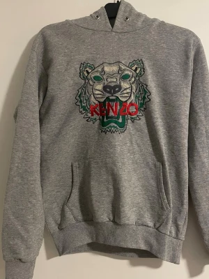 Grå Kenzo hoodie med tigerbrodyr - Snygg grå hoodie från Kenzo med ikonisk tigerbrodyr och röd Kenzo-logga på bröstet. Hoodien har huva, magficka och ribbade muddar. Perfekt för dig som vill sticka ut med en streetstyle-look.