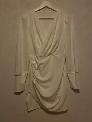 Vit klänning - Stilren cremevit kortklänning med omlott framtill och snygg drapering i kjolen. Klänningen har långa ärmar med breda manschetter och v-ringad urringning. Perfekt för dig som gillar minimalistisk och elegant stil. Använd 1 gång!
