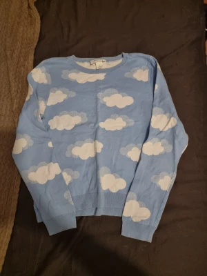 Blå stickad tröja med moln från Monki - Superfin blå stickad tröja från Monki med vita moln över hela. Tröjan har rund halsringning, långa ärmar och ribbade muddar vid ärmslut och nederkant. Perfekt för dig som gillar lekfulla och unika plagg.