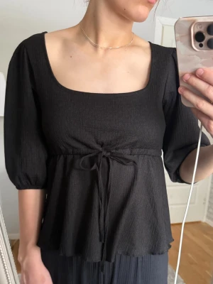 Svart topp med knyt från 157 - Svart topp från 157 med fyrkantig ringning, puffiga trekvartsärmar och knyt i midjan. Tunt, lätt material som ger ett flowy fall. Perfekt för dig som gillar en enkel men ändå trendig look.