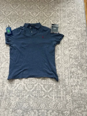 Blå pikétröja från Ralph Lauren - Snygg blå pikétröja från Ralph Lauren med klassisk krage och knappar framtill. Tröjan har det ikoniska röda Polo-logotypen broderad på bröstet. Perfekt till jeans eller shorts för en avslappnad men stilren look.