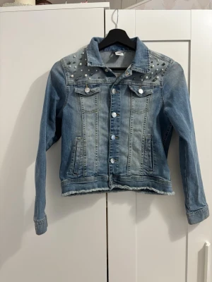 Ljusblå jeansjacka med pärlor - Trendig ljusblå jeansjacka med pärldetaljer på axlarna och rå kant nertill. Klassisk knappstängning framtill, två bröstfickor med lock och två sidofickor. Perfekt för dig som vill ha en cool och unik look. Skriv gärna innan ni köper❤️