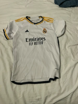 Real Madrid Benzema matchtröja - Säljer en vit Real Madrid matchtröja från Adidas med gula och svarta detaljer. Tröjan har klubbmärke och sponsortryck fram samt Benzema och nummer 9 på ryggen. Tillverkad i lätt och ventilerande material, perfekt för fotbollsträning eller match.