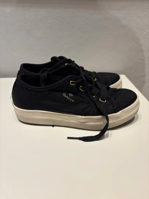 Svarta låga sneakers från Gant - Stilrena svarta sneakers från Gant med vita sulor och guldfärgade detaljer. Skorna är låga och har klassisk snörning samt diskret logga på sidan. Skorna är sparsamt använda så fint skick!! 