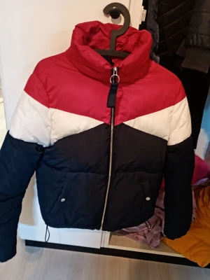 Färgglad pufferjacka från Bershka - Snygg kort pufferjacka från Bershka i färgblock med rött upptill, vitt i mitten och mörkblått nedtill. Jackan har hög krage, dragkedja framtill och två fickor med knappar. 