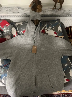 Burrbery kofta  - En hoodie från Burberry i ljusgrått. Den har dragkedja framtill, huva med snörning och stora fickor. Insidan av huvan har ett klassiskt Burberry-mönster. Perfekt för dig som gillar stilrena och bekväma plagg med exklusiv känsla. 500kr 