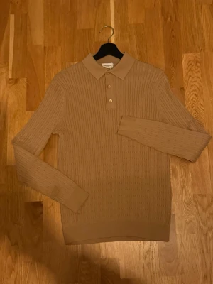 Beige stickad tröja från John Henric - Stickad tröja i beige från John Henric med klassisk kabelstickning, krage och tre knappar framtill. Oanvänd med prislappar. Tröjan har lång ärm och ribbade muddar vid ärmslut och nederkant. Perfekt för en stilren och avslappnad look. Pris kan diskuteras