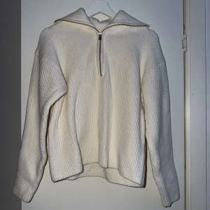 Vit stickad tröja med halv dragkedja - Säljer en krämvit stickad tröja med ribbad struktur och halv dragkedja framtill. Tröjan har en bred krage och avslappnad passform med långa ärmar. Perfekt för lager på lager och en clean look.