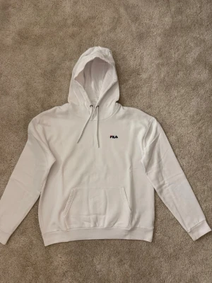 Vit hoodie från FILA  - Säljer en stilren vit hoodie från FILA med liten broderad logga på bröstet. Hoodien har klassisk känguruficka, dragsko i huvan och ribbade muddar vid ärmar och nederkant. 