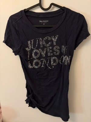Juicy Couture mörkblå t-shirt - Mörkblå t-shirt från Juicy Couture med glittrig text 'Juicy Loves London' framtill. T-shirten har korta ärmar och en knytdetalj i sidan. Tröjan är använd men i hyfsat bra skick!