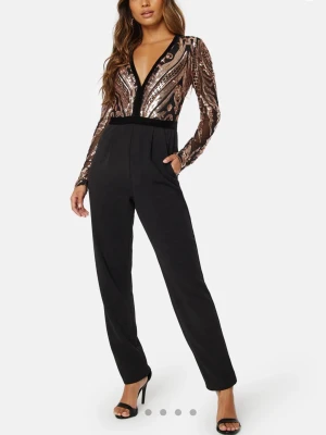 Svart jumpsuit med paljettöverdel - Snygg svart jumpsuit med långärmad överdel täckt av bronsfärgade paljetter och djup V-ringning. Byxdelen är enkel och rak med fickor, medan överdelen har transparenta meshpartier och coolt mönster. Perfekt för dig som vill sticka ut. Från bubbelroom, helt slutsåld och använd en gång. 