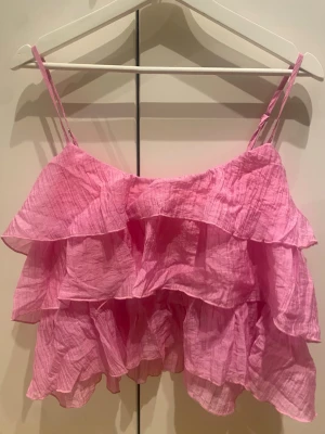 Rosa volanglinne med tunna axelband - Supersött rosa linne med flera volanger och tunna axelband. Toppen är gjord i ett lätt, krinklat tyg som ger en luftig känsla. Perfekt för sommardagar och att styla med jeans eller kjol. Köptes på sellpu ingen aning på märken 