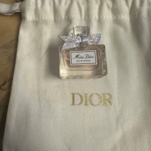 Miss Dior Eau de Parfum - Säljer en elegant flaska Miss Dior Eau de Parfum. Flaskan är liten och rektangulär med en söt rosett på korken. Parfymen har en ljusrosa nyans och kommer i en fin vit påse med Dior-loggan i guld. Perfekt för att ge en lyxig touch till din doftsamling!
