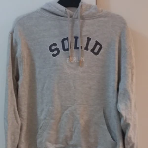 Grå hoodie från Solid Berlin - Säljer en grå hoodie från Solid Berlin i bra skick. Den har en klassisk design med en stor ficka fram och justerbar huva med snören. Perfekt för en avslappnad stil och passar till alla årstider. Skön och enkel att matcha med jeans eller joggers.