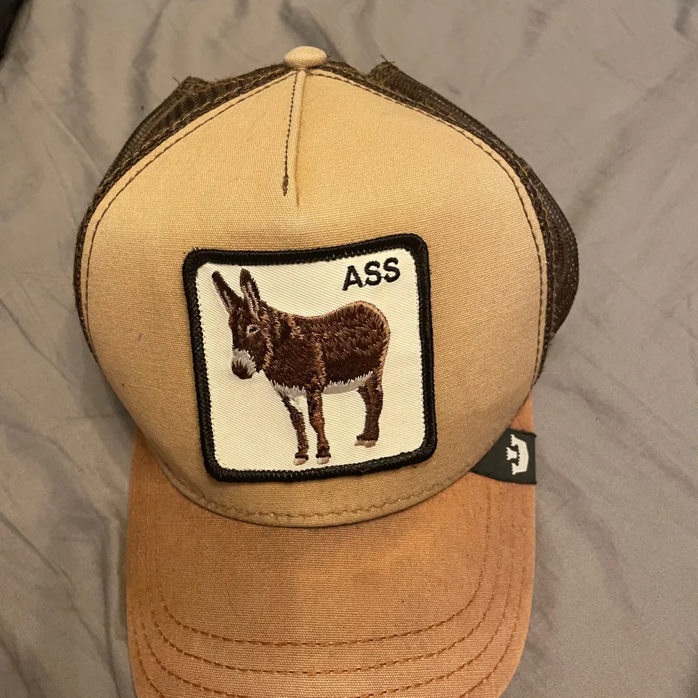Säljer en beige och brun keps från The Farm med ett broderat märke av en åsna på framsidan. Kepsen har ett nätmaterial baktill för bra ventilation och en justerbar rem för perfekt passform. Perfekt för en avslappnad stil eller en dag på landet! 🐴. Asusteet.