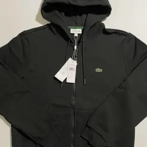 Säljer en svart hoodie från Lacoste i nytt skick. Den har en dragkedja framtill och en klassisk krokodil-logga på bröstet. Perfekt för höst och vår, med långa ärmar och en skön huva. Passar både till vardags och chill.