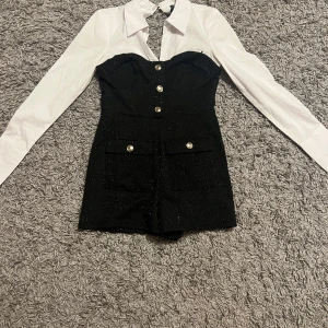 Svart glittrig byxdress - Snygg svart glittrig byxdress med vita långa ärmar och knappar framtill. Den har en tight passform och är perfekt för festliga tillfällen. Klänningen har en dragkedja bak och dekorativa fickor framtill. Perfekt för en kväll ute eller en speciell tillställning! Från Newyorker