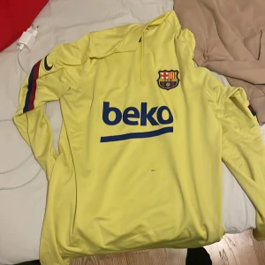 Gul FC Barcelona träningsjacka - Säljer en gul FC Barcelona träningsjacka från Nike. Den har långa ärmar med ränder i rött och blått på sidorna. Jackan har en halv dragkedja framtill och klubbens emblem på bröstet. Perfekt för fotbollsträning eller som en snygg vardagsjacka!