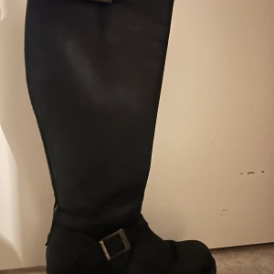 Johnny Bulls - Snygga svarta stövlar i skinn med ett coolt spänne vid ankeln. Perfekta för höst och vinter, dessa stövlar är både stiliga och praktiska. De har en dragkedja på sidan för enkel på- och avtagning. Storlek 40 och i nytt skick. Perfekta för att ge din outfit en edgy touch!