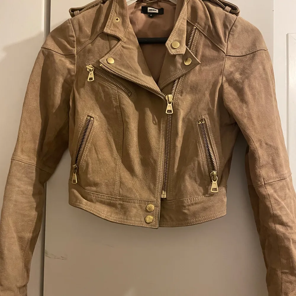 Snygg beige/brun mockajacka från bikbok i mycket bra skick. Jackan har en bikerstil med dragkedjor och knappar i guld. Perfekt för höst och vår. Den är croppad och har långa ärmar. Storlek S . Jackor.