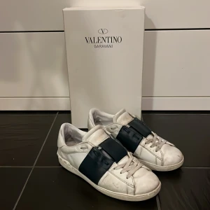 Valentino Garavani sneakers - Säljer ett par snygga Valentino Garavani sneakers i vitt med breda mörkblå detaljer. Skorna har en stilren design med snörning och är i bra skick. Perfekta för både vardag och festliga tillfällen. Kommer med originalkartong.